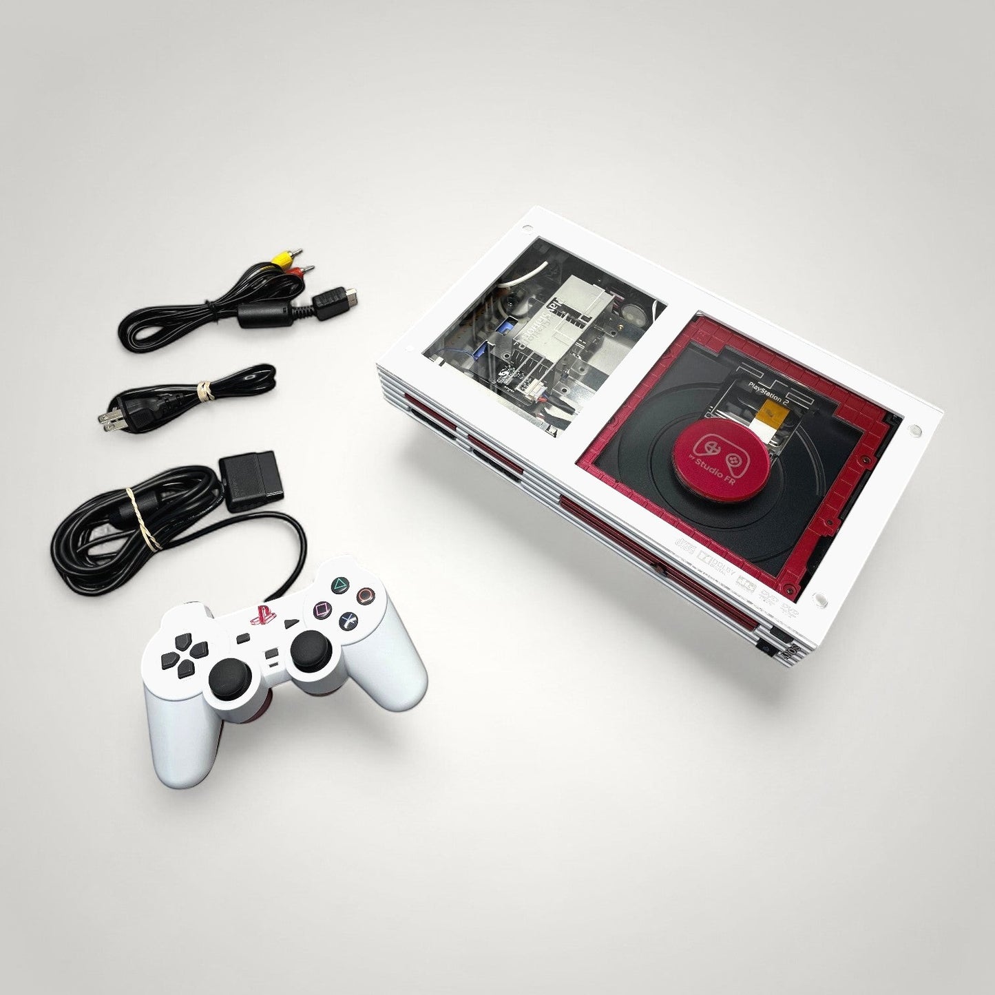 StudioFR custom red and white PlayStation 2 Fat console with clear top, controller, and cables.