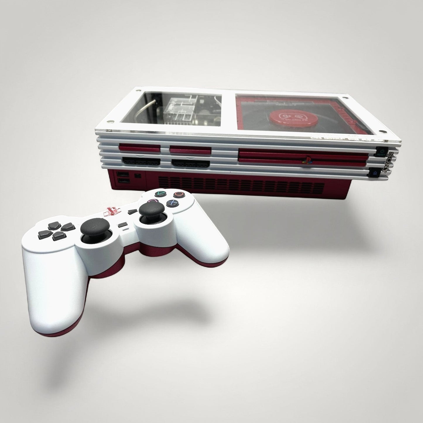 StudioFR custom red and white PlayStation 2 Fat console with matching controller. Features clear top with visible internals and RGB lighting.